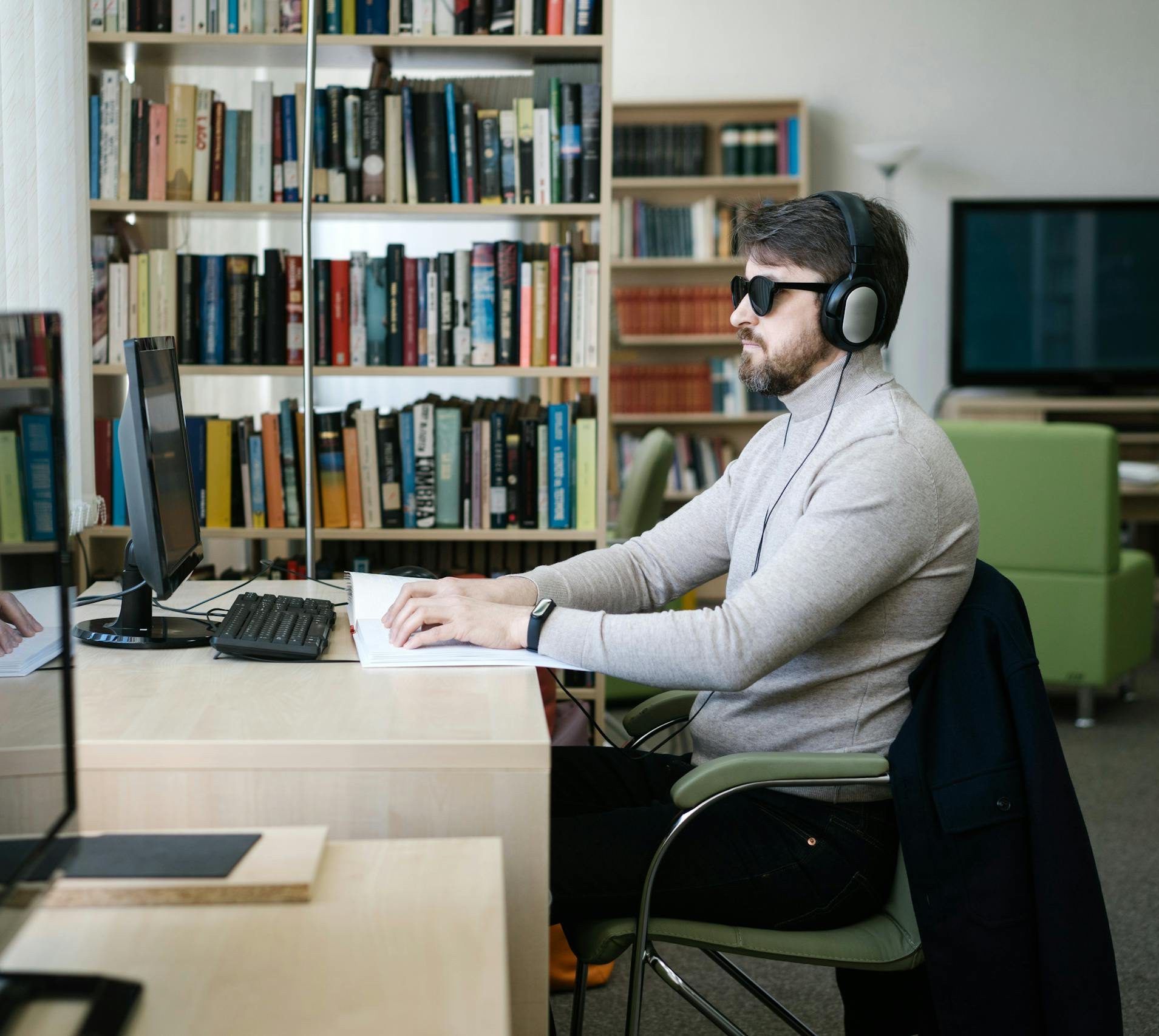 A blind man uses a computer with headphones in a library, highlighting technology's role in accessibility.